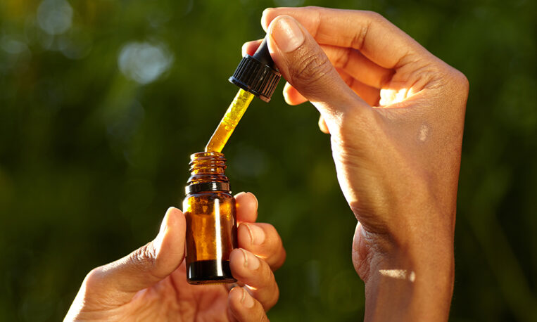 Close-up of hands holding an amber glass dropper bottle, lifting a dropper of golden oil against a blurred green background.