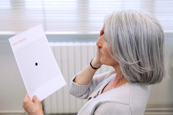 A senior woman with gray hair holds an Amsler grid at eye level, covering one eye to perform a self-vision test.